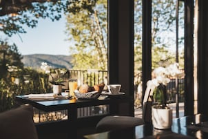 Uma mesa com um prato de croissants, uma chávena e um copo de sumo de laranja, com vista para árvores e montanhas através de grandes janelas.