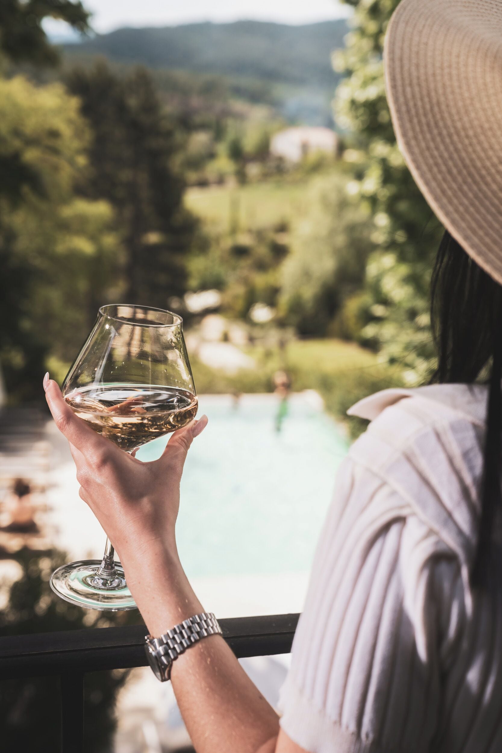 A person holding a wine glass with a view of a swimming pool and greenery in the background.