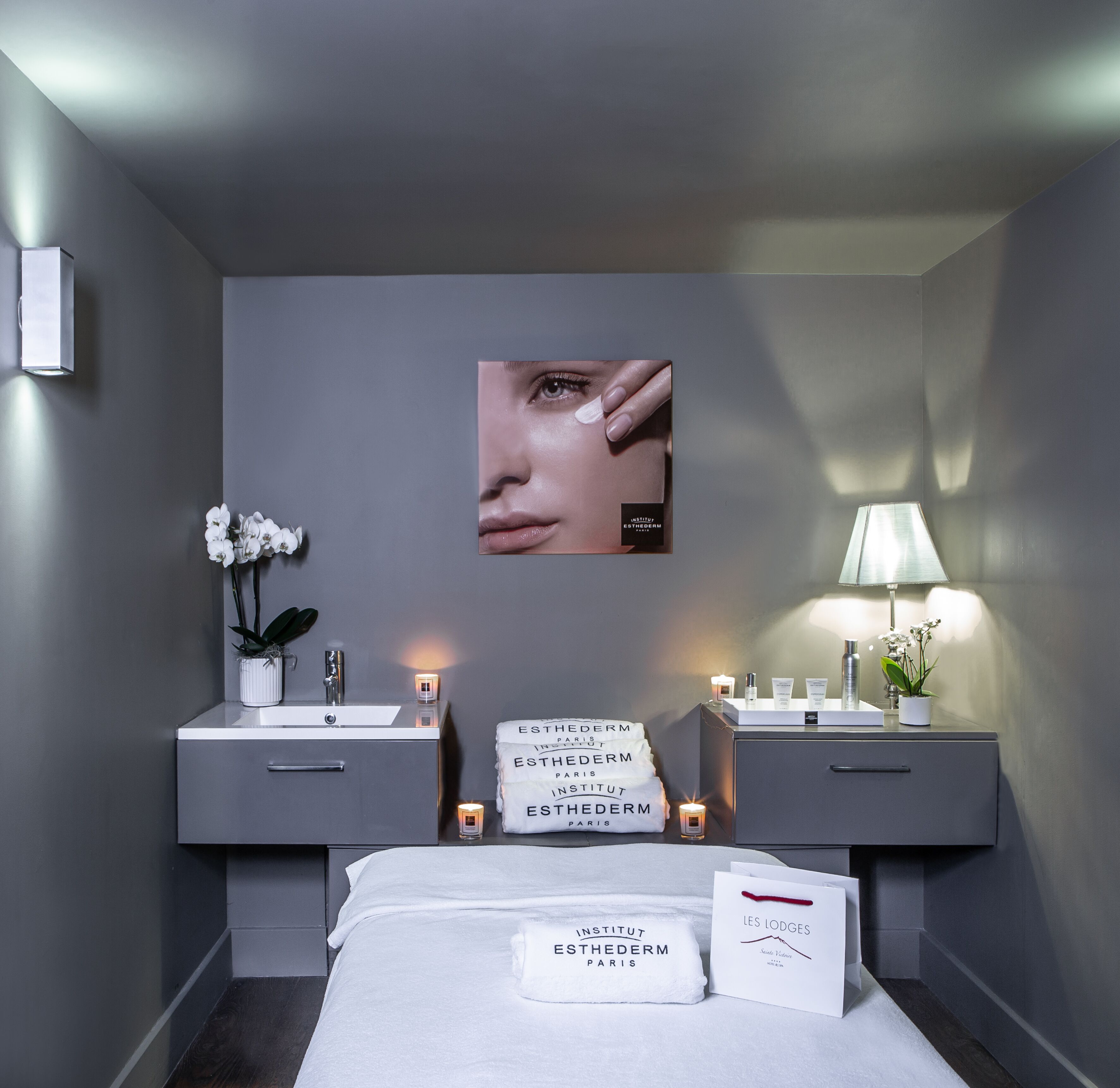Une chambre d’hôtel moderne aux tons de gris, avec un grand miroir, deux vasques, un lit recouvert d’une couette blanche et un tableau encadré au mur.