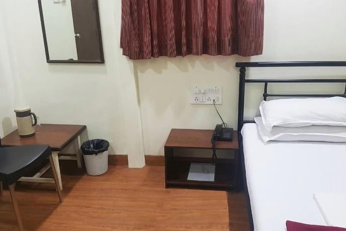 A small, tidy room with a bed, a desk, a chair, a trash bin, and a wall-mounted mirror.
