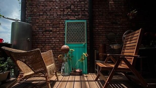Una terraza de madera con sillas de mimbre y una mesa de madera, una puerta verde y un muro de ladrillo.