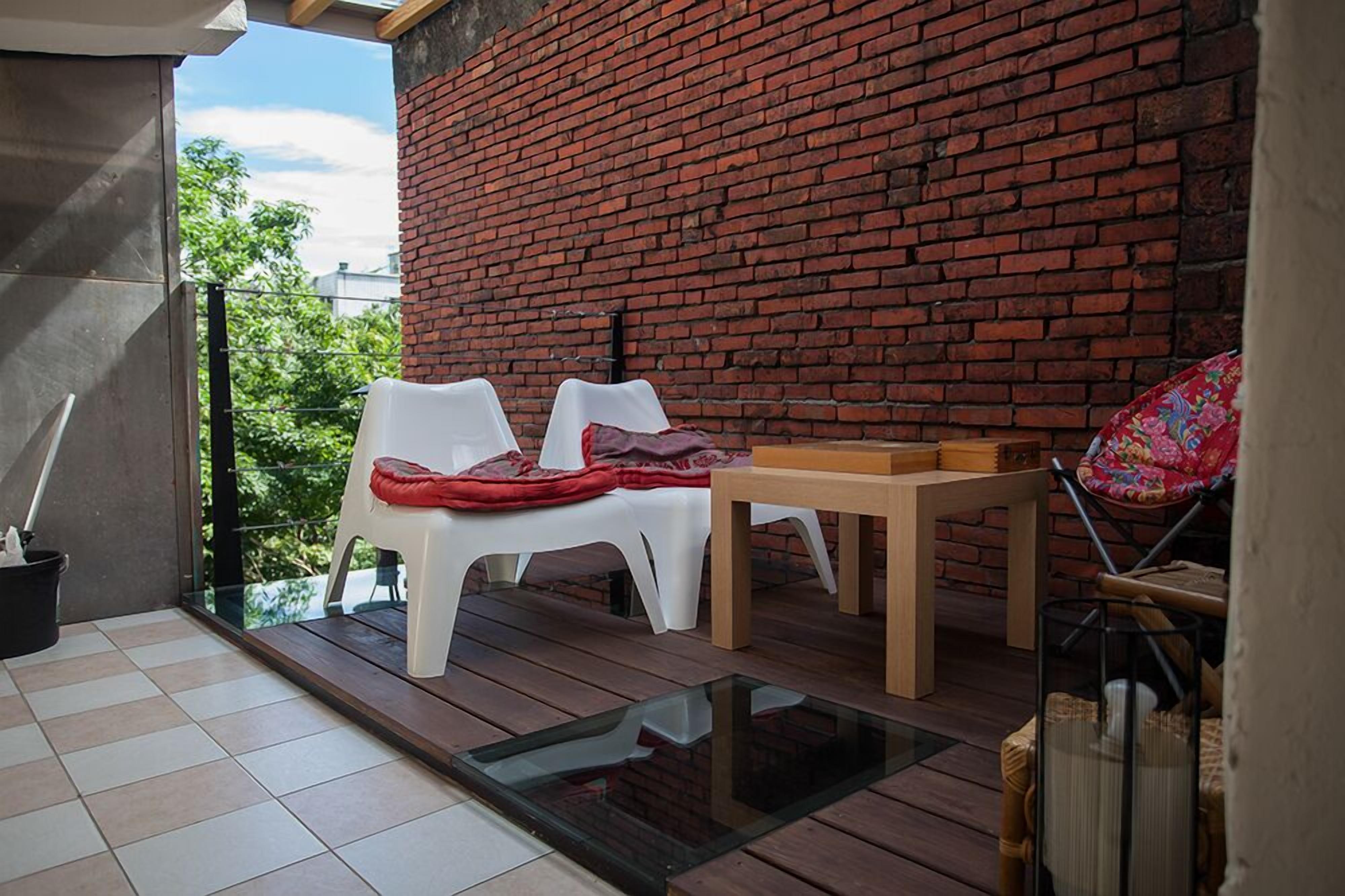 A balcony with a brick wall, two white plastic chairs, a wooden table, and a wicker chair.