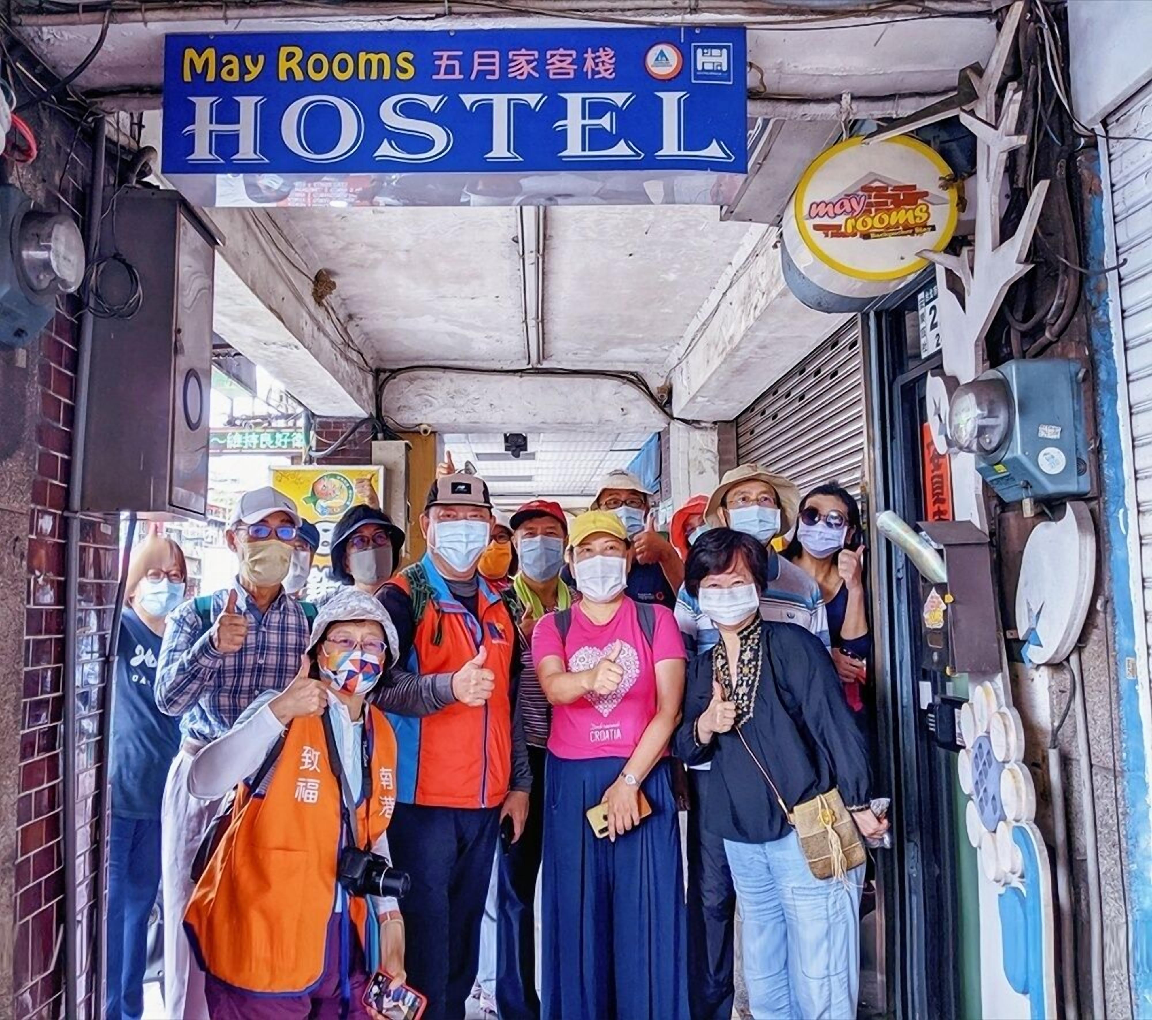 Un grupo de personas de pie frente a un albergue con un letrero que dice "May Rooms" y "HOSTEL".