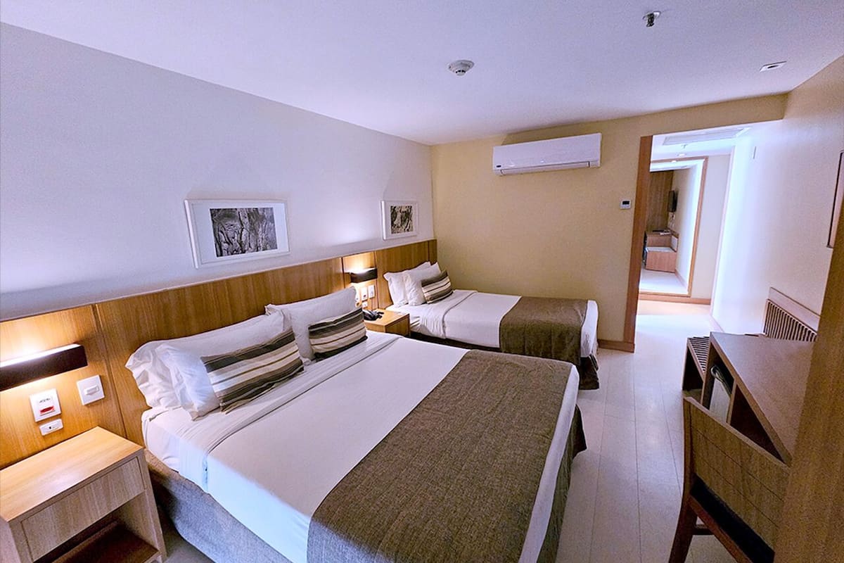A hotel room with two beds, a desk, and a chair. There are wall-mounted air conditioning units and framed pictures on the walls.