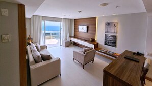 A modern living room with a sofa, armchair, and a wooden coffee table. There is a TV mounted on the wall and a window with curtains.