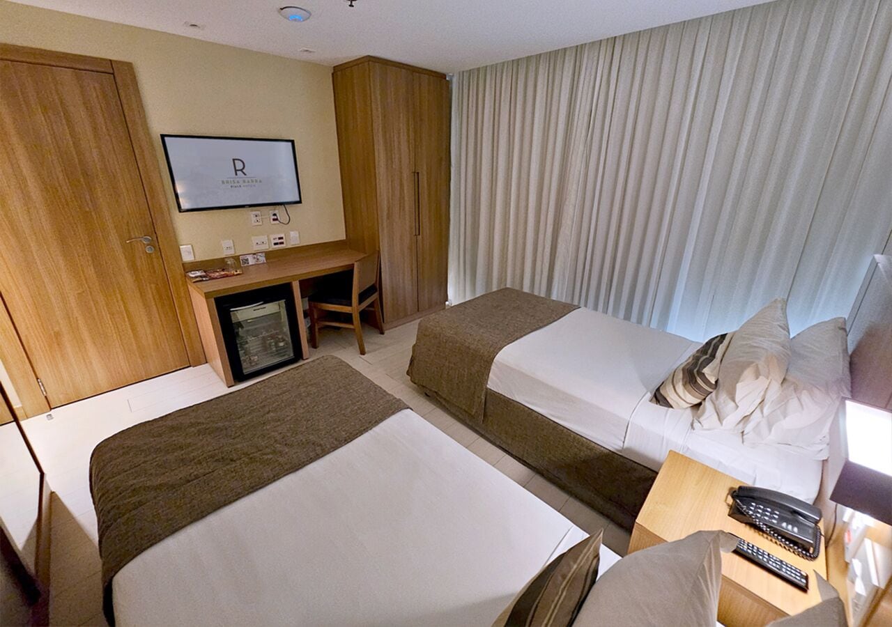 Habitación de hotel con dos camas, un escritorio con computadora, un televisor en la pared y una ventana con cortinas.