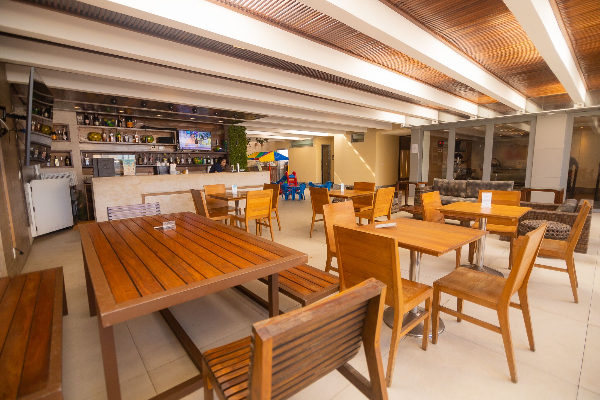 Un restaurante con mesas y sillas de madera, una zona de bar con estantes llenos de botellas y una pantalla de televisión.