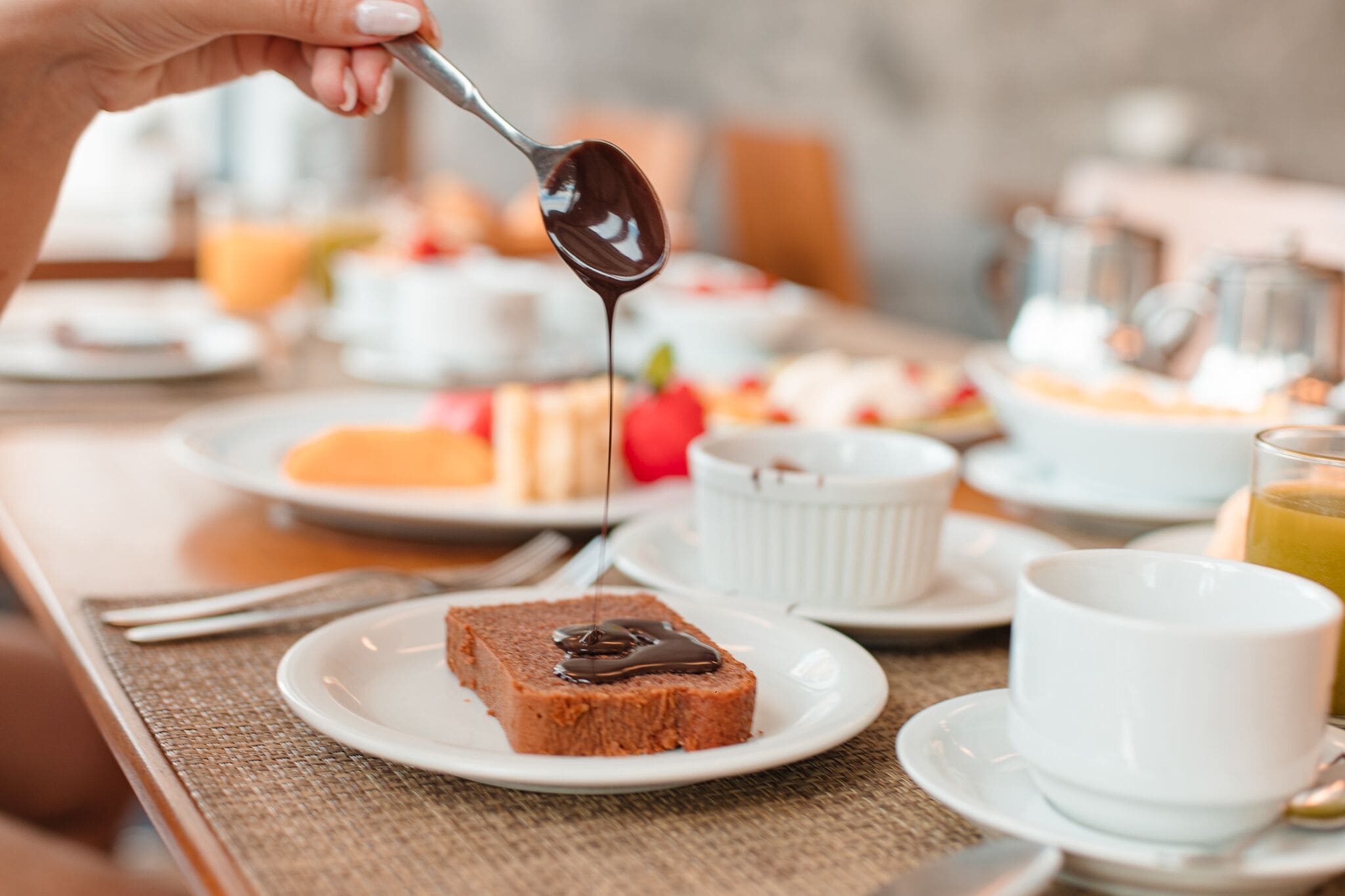 A breakfast setting with a slice of cake topped with chocolate sauce on a plate, a white cup, and a glass of orange juice.