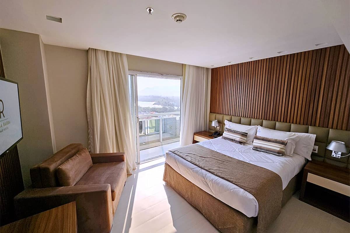 A modern hotel room with a large bed, a sofa, a desk, and a view of the city through the window.