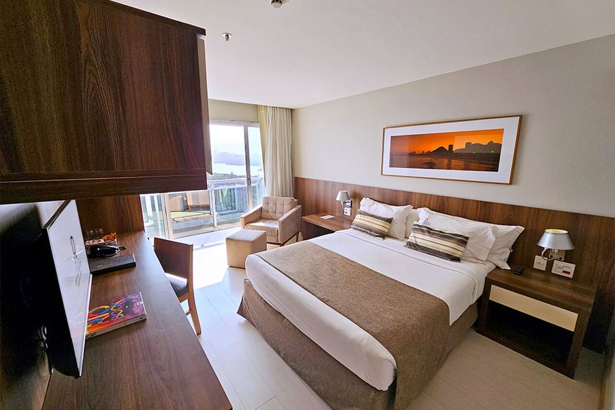 A hotel room with a large bed, a desk with a computer, a chair, a TV, and a window with a city view.