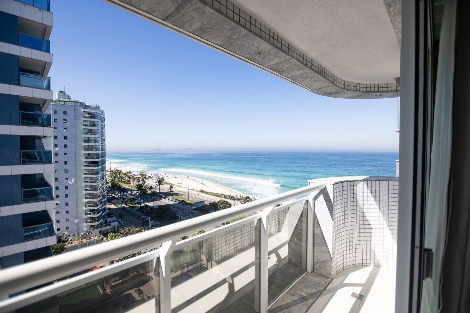 Un balcon offrant une vue sur une plage et l’océan, donnant également sur un parking et un immeuble de grande hauteur.