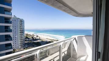 Un balcon offrant une vue sur une plage et l’océan, donnant également sur un parking et un immeuble de grande hauteur.