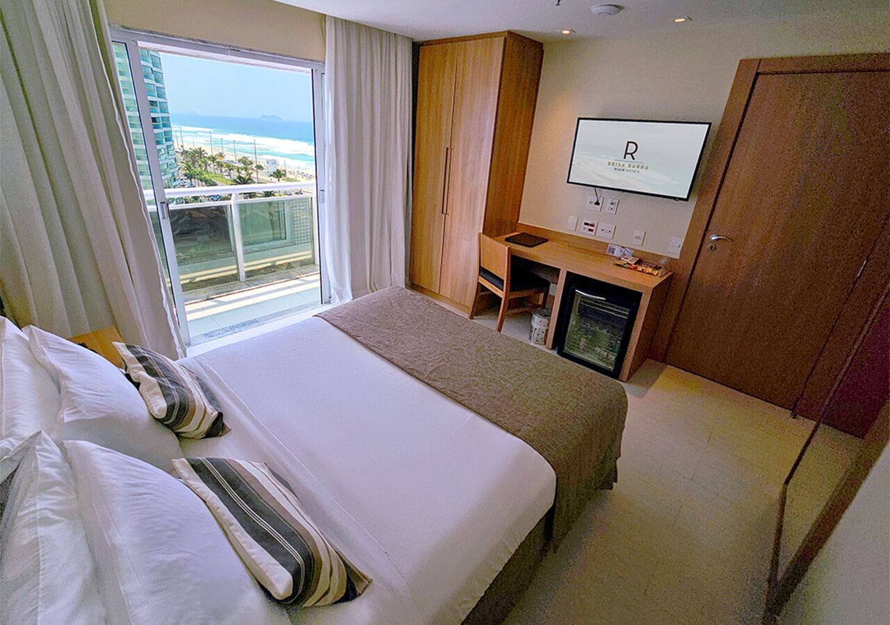 A hotel room with a bed, a desk, a television, and a view of the ocean.