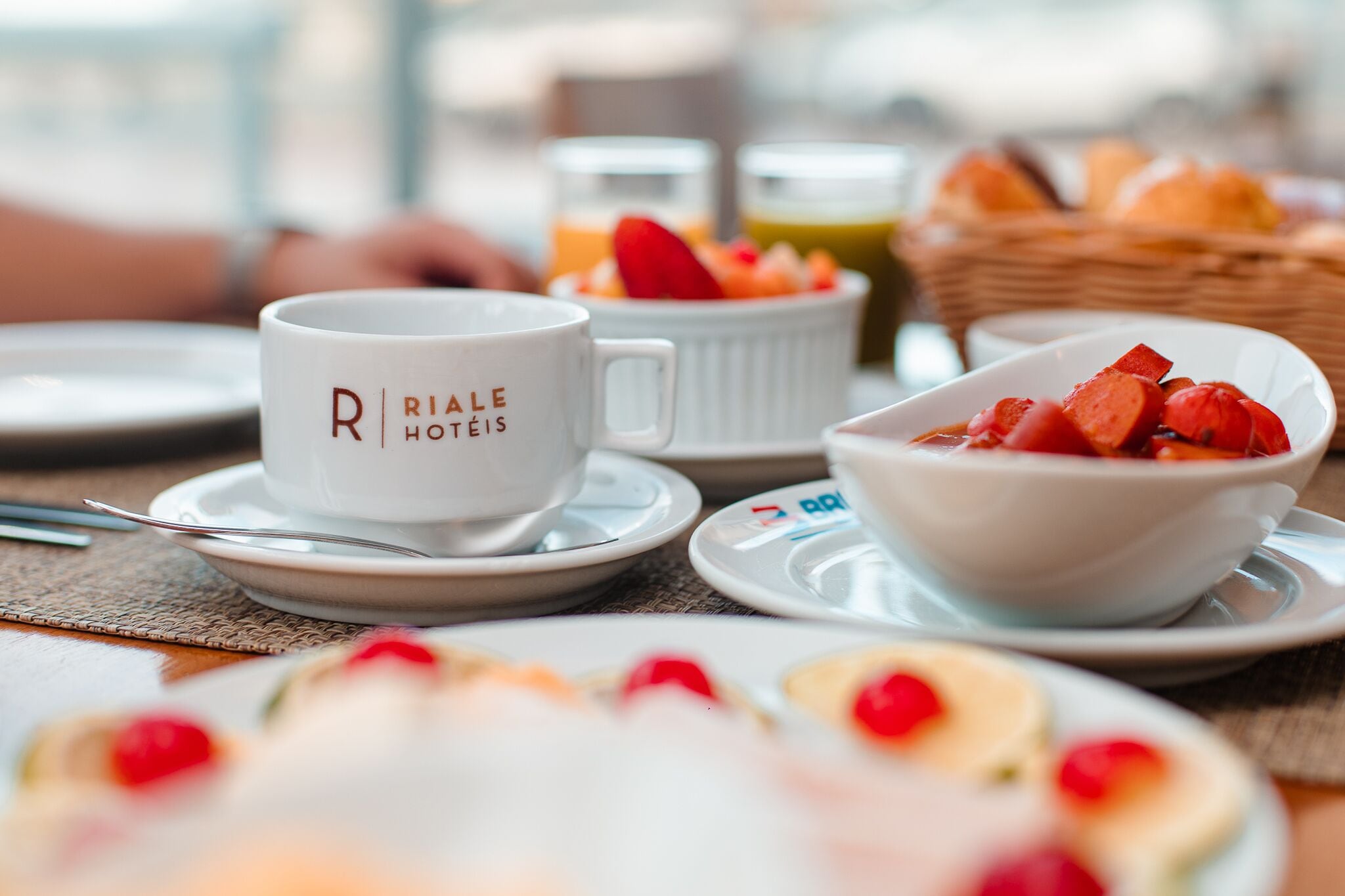 A breakfast setting with a white mug labeled "Rialle Hotels," a bowl of fruit, and a basket of bread.