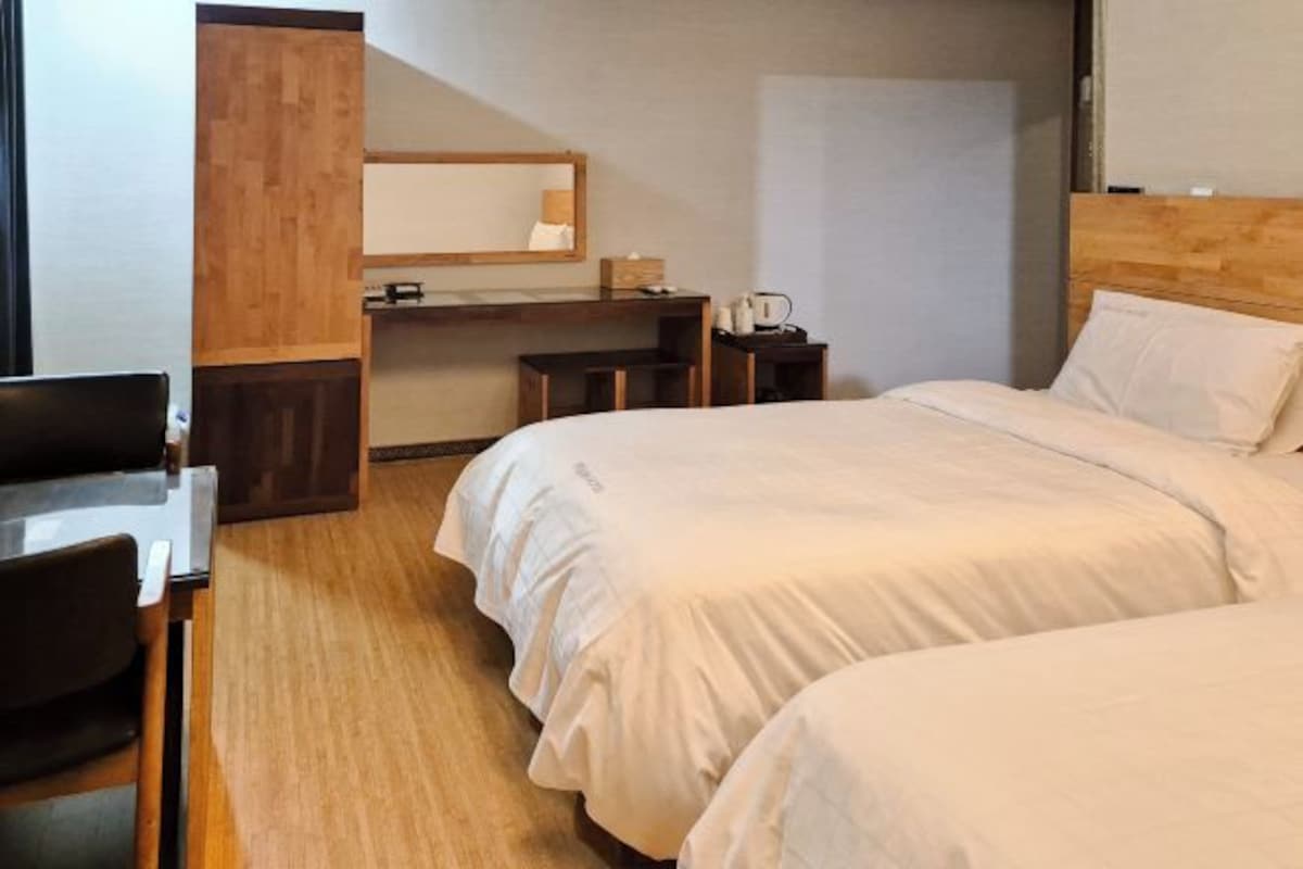 A hotel room with two beds, a wooden desk, a mirror, and a small table with a kettle.