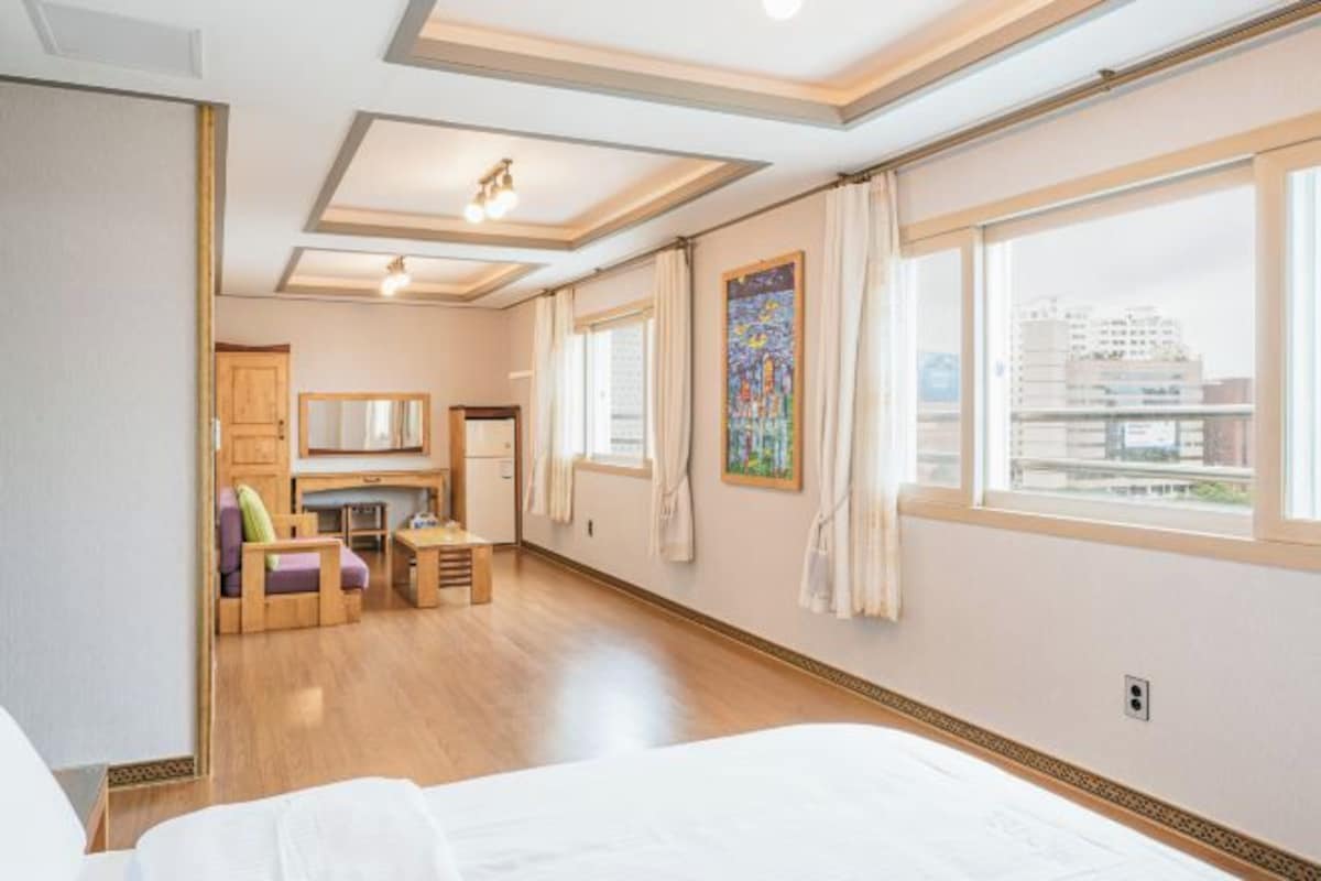 A hotel room with a bed, a desk, a chair, a window with a city view, and a painting on the wall.
