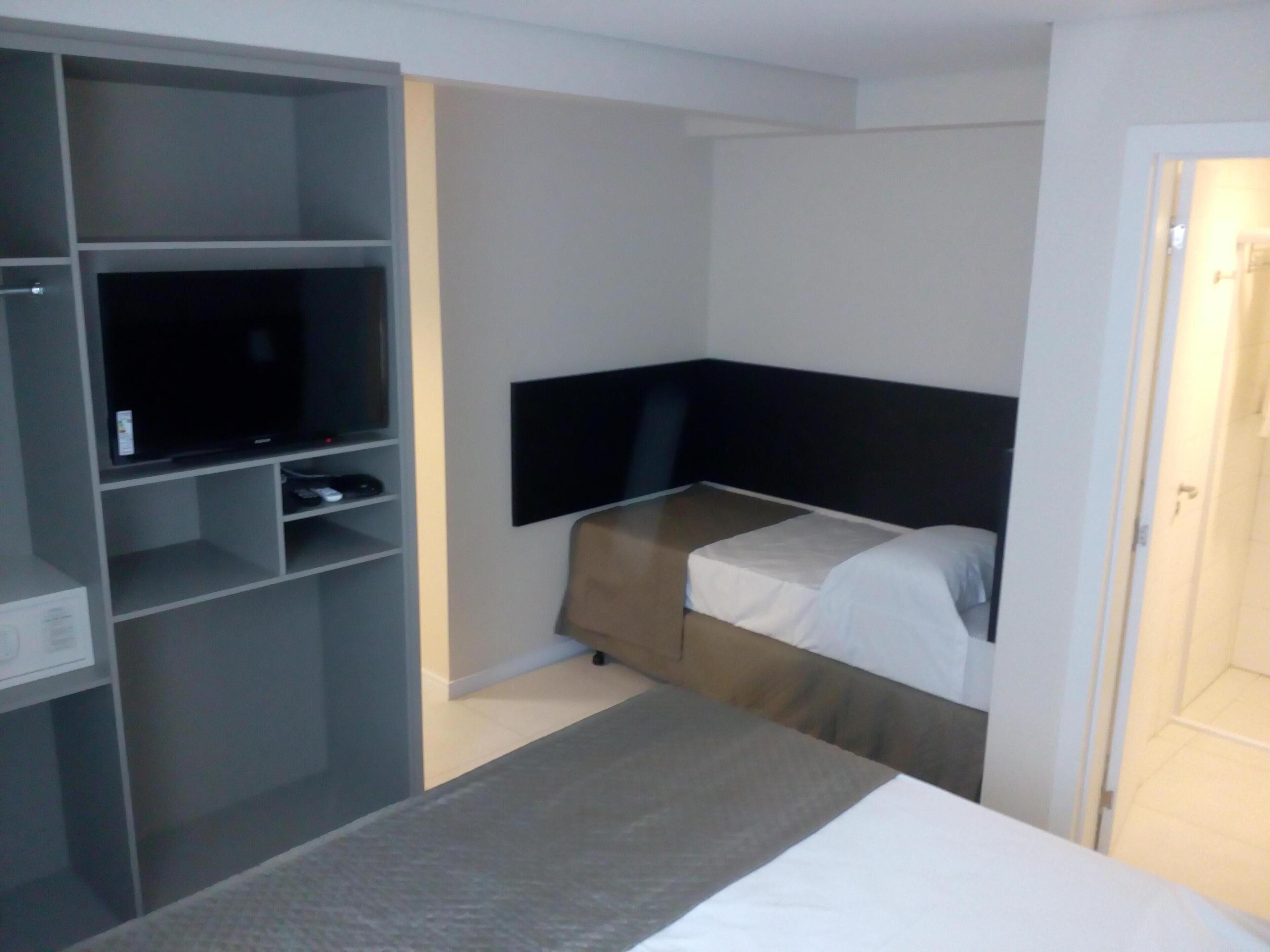 Een kleine hotelkamer met een bed, een televisie op een plank en een kledingkast.