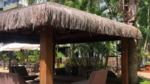 Un área de estar al aire libre con techo de paja, sillones reclinables y una mesa, rodeada de palmeras y un edificio al fondo.