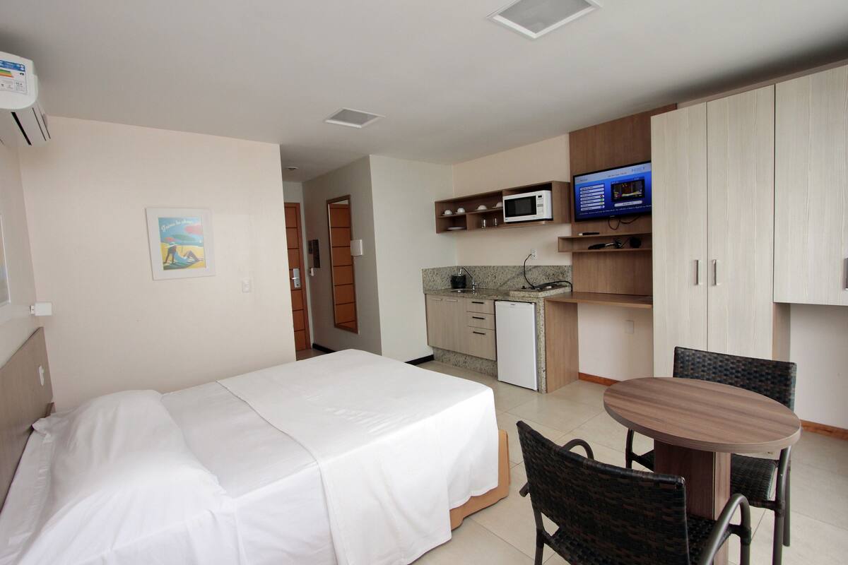 Habitación de hotel con cama, kitchenette pequeña, mesa de comedor con sillas y televisión en la pared.