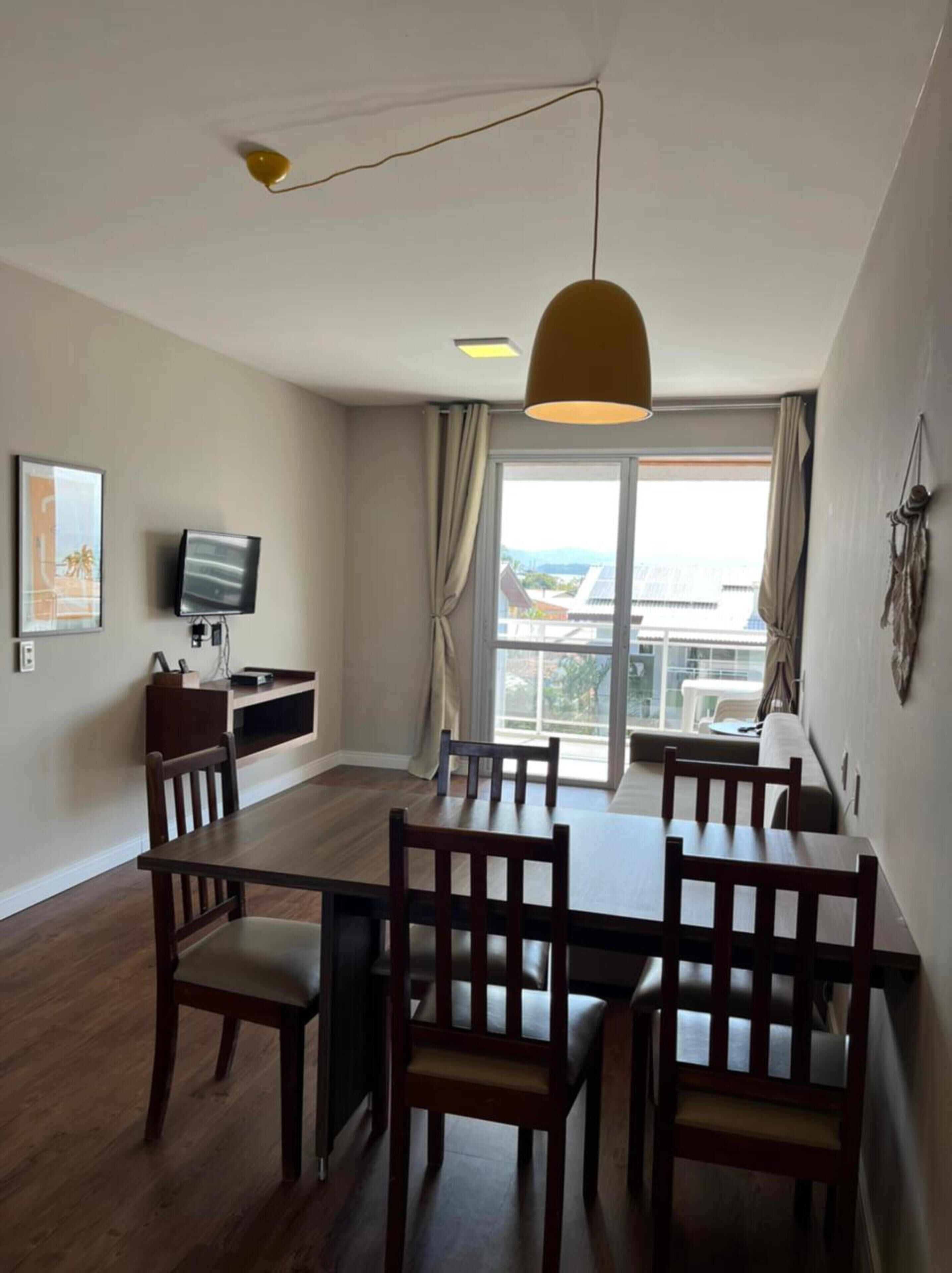 A modern dining area with a wooden table and chairs, a television on the wall, and a large window with curtains.