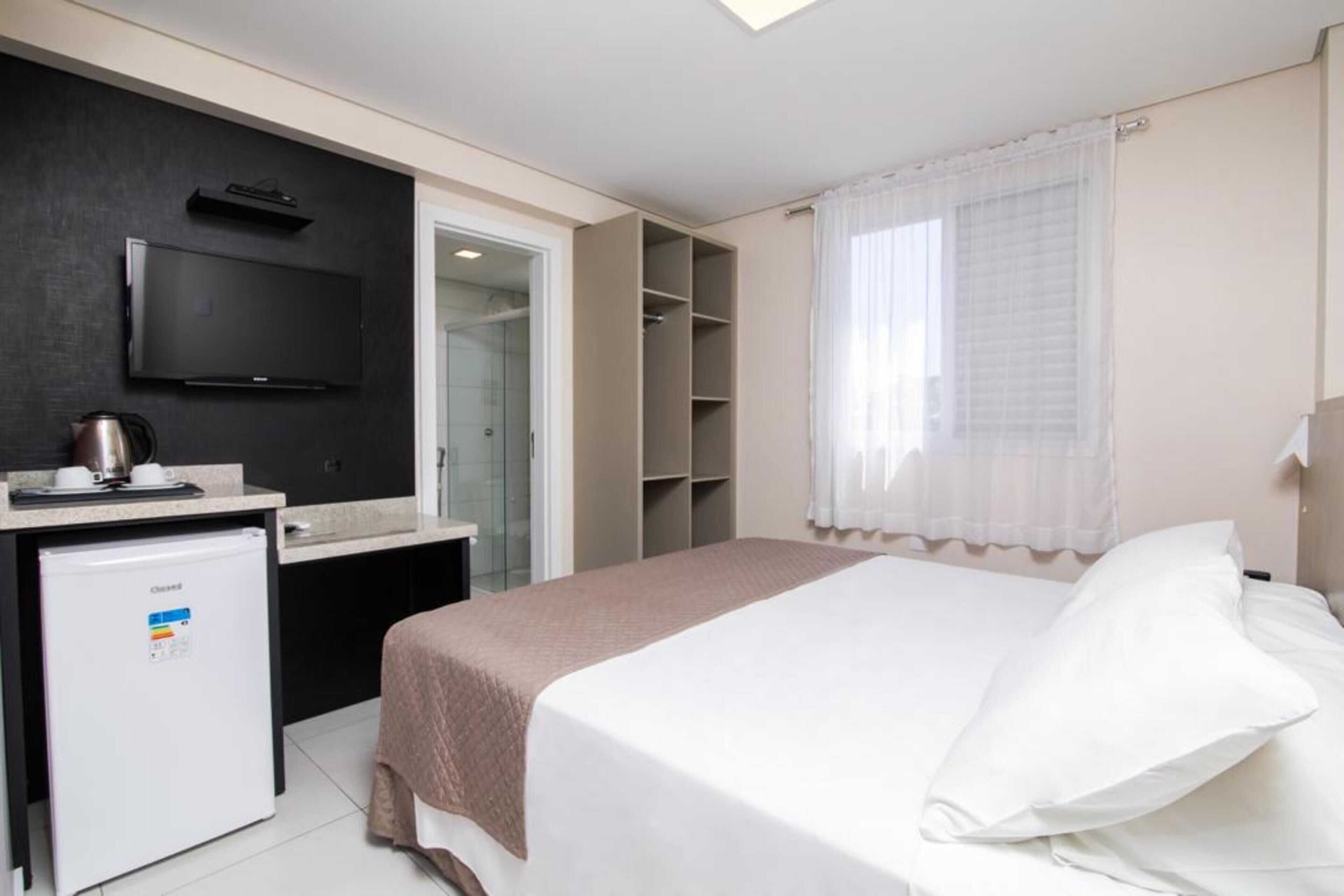 Habitación de hotel con cama, televisor montado en la pared, pequeña kitchenette con refrigerador y vestidor.