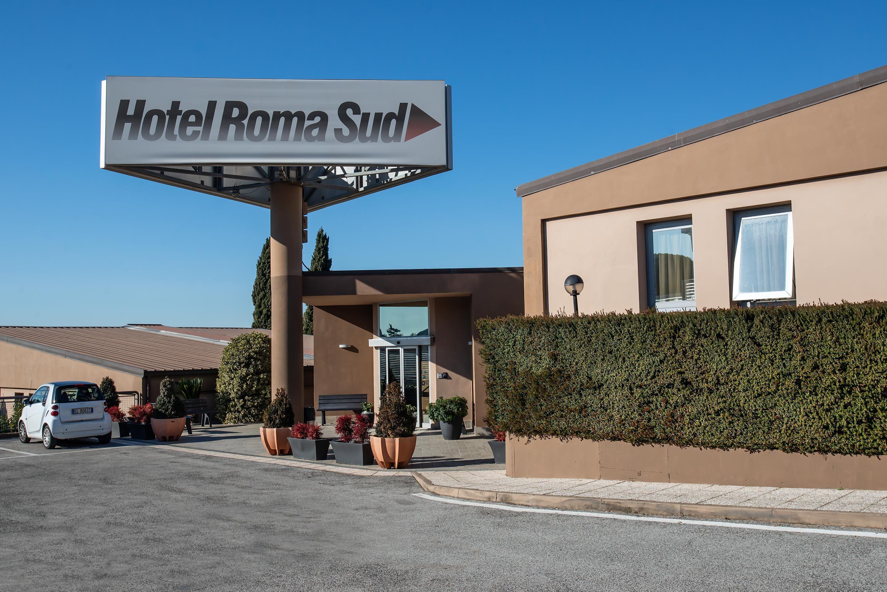 L’entrée d’un hôtel, avec une enseigne indiquant « Hôtel Roma Sud », et un espace de stationnement où se trouve une voiture blanche.