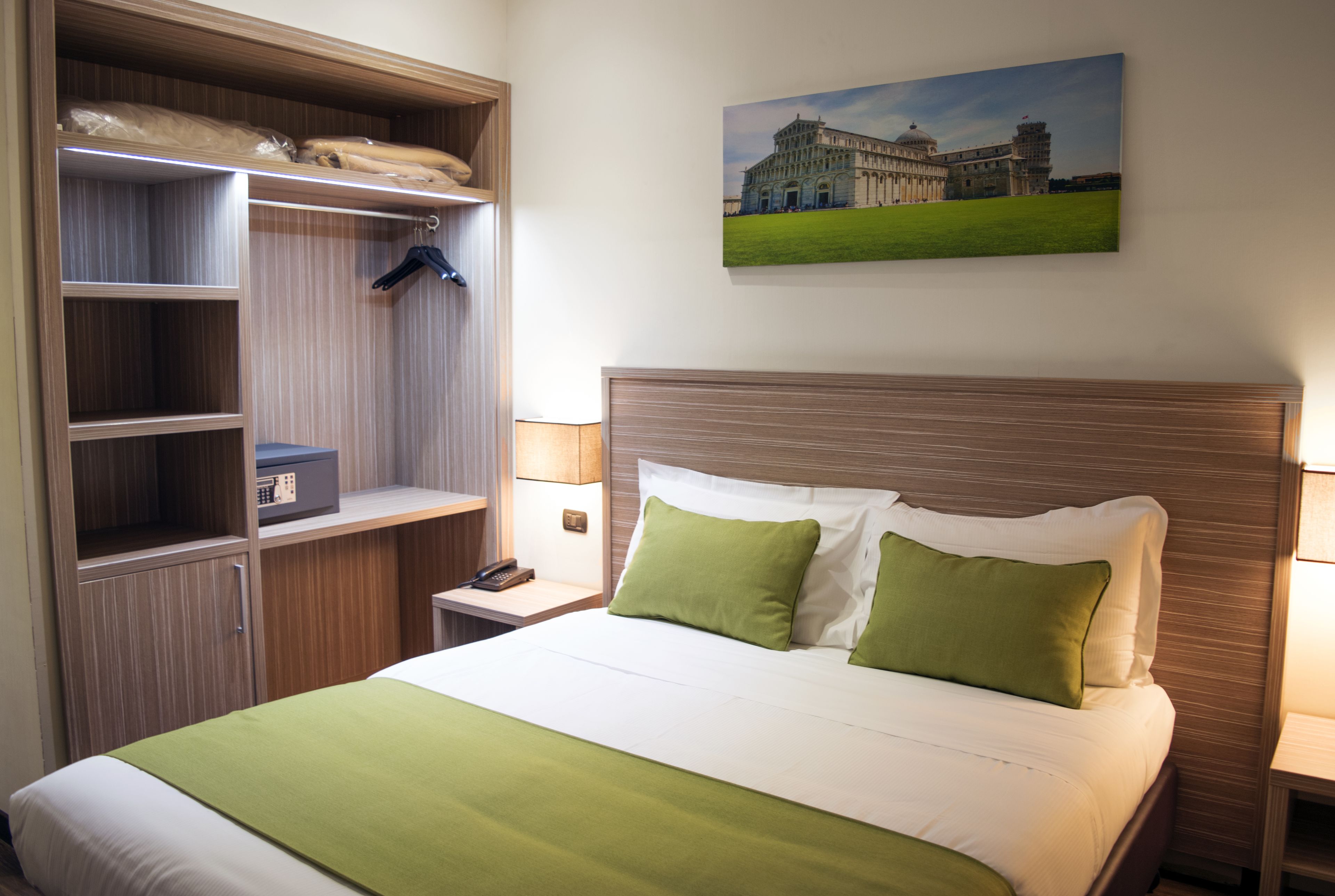 Habitación de hotel con una cama bien hecha, un armario con ropa colgada, una mesita de noche con lámpara y un cuadro de un edificio en la pared.