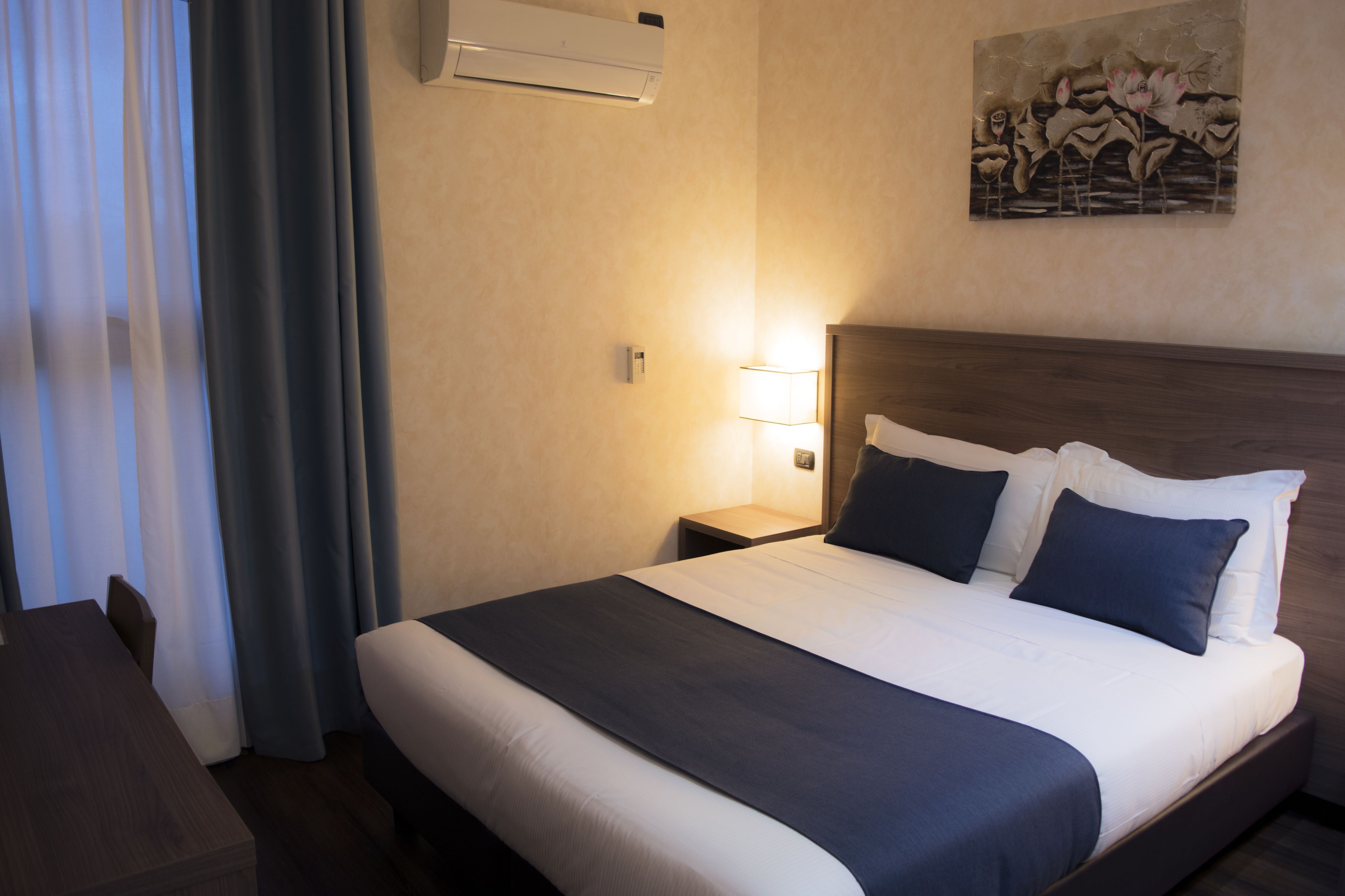 A hotel room with a large bed, a bedside table with a lamp, a wall-mounted air conditioner, and a painting on the wall.