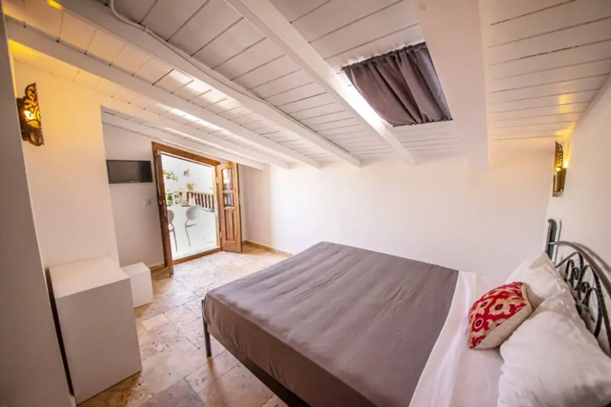 A bedroom with a bed, a small bedside table, a wardrobe, a ceiling fan, and a door leading to a balcony.