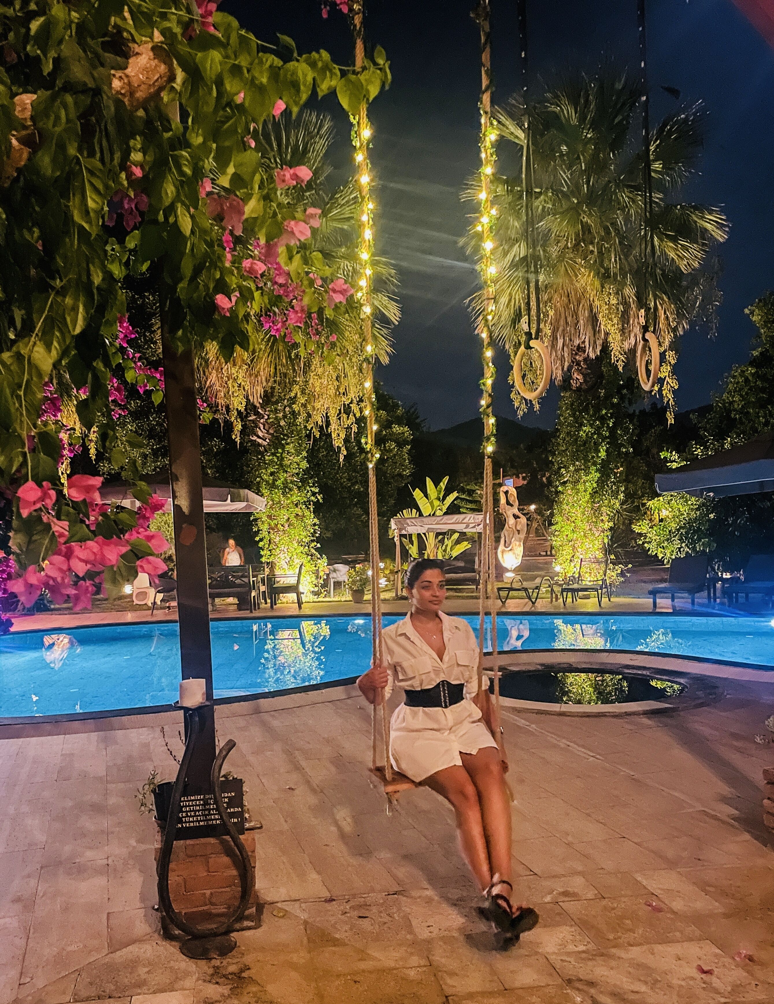 A woman sitting on a swing in a well-lit outdoor area with a pool, surrounded by greenery and decorative lights.