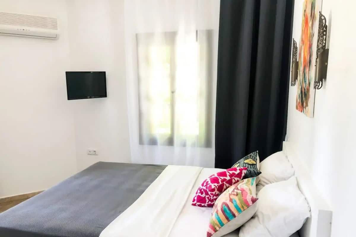 A bedroom with a bed, a flat-screen TV, a window with curtains, and a wall-mounted air conditioner.