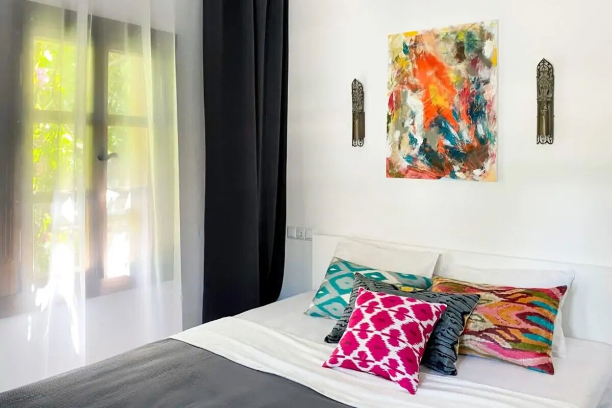 Un dormitorio con una cama, una ventana con cortinas traslúcidas y un cuadro abstracto de colores en la pared.