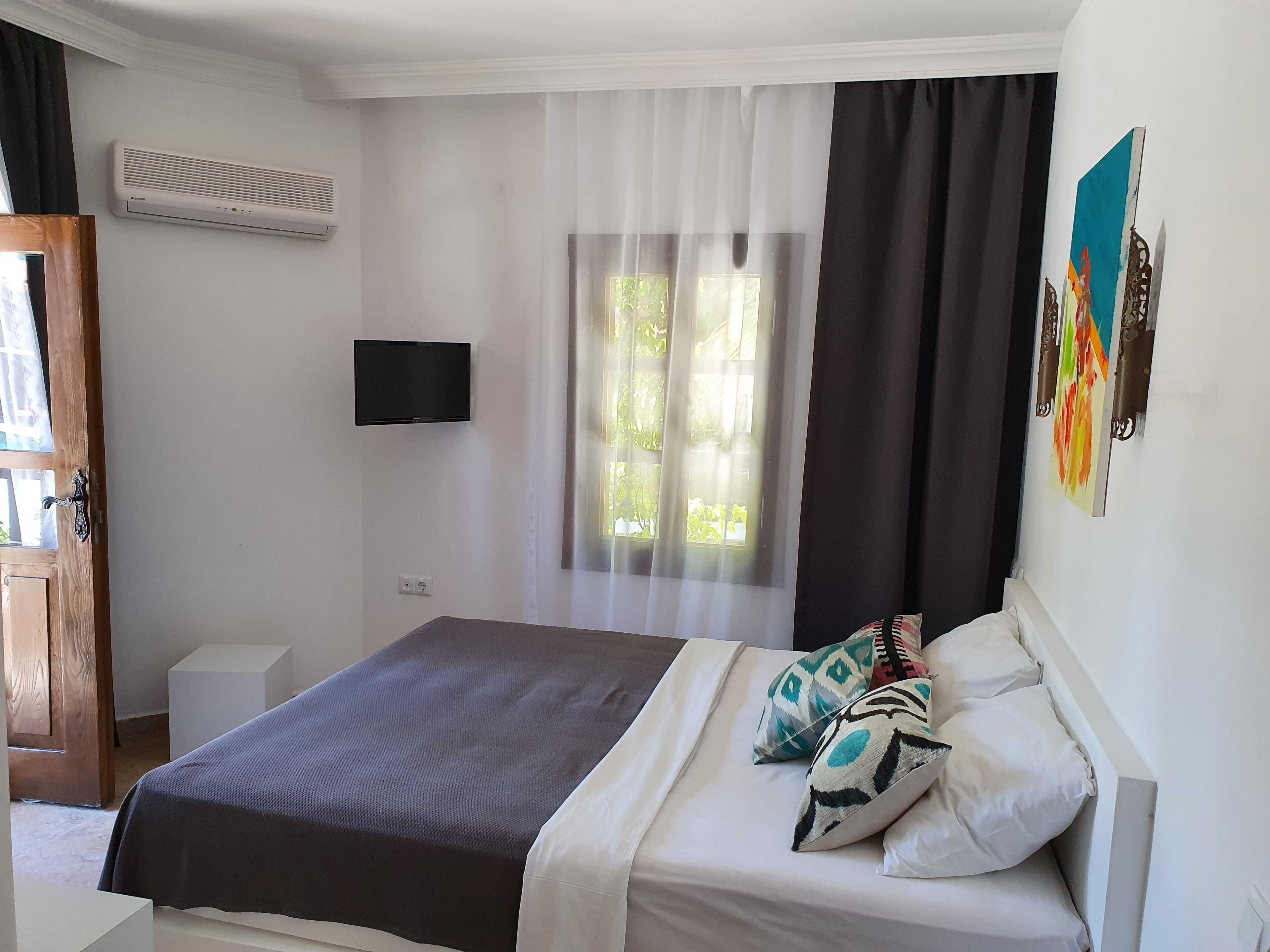 A bedroom with a bed, a TV mounted on the wall, a window with curtains, and a door.