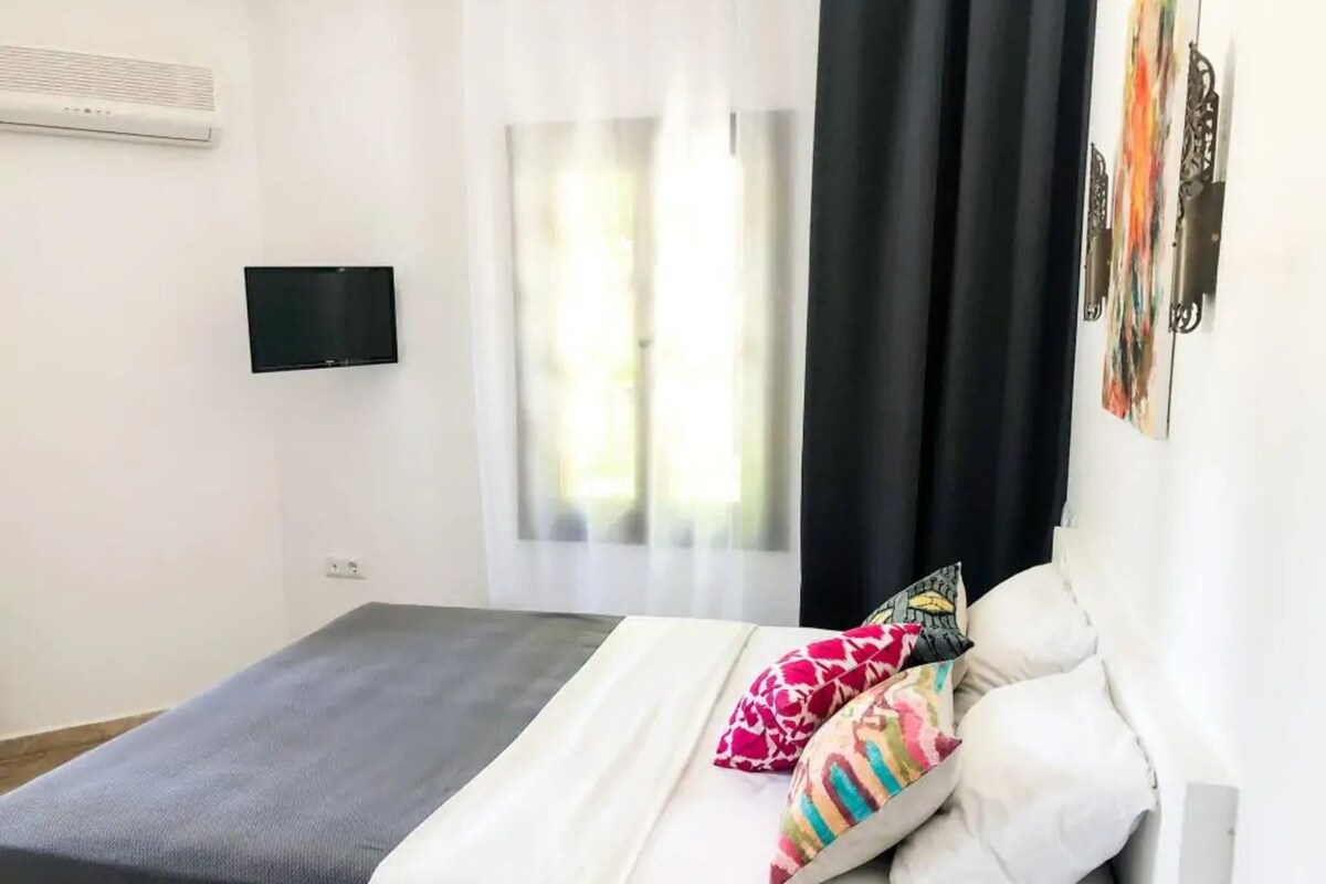A bedroom with a bed, a flat-screen TV, a window with curtains, and a wall-mounted air conditioner.