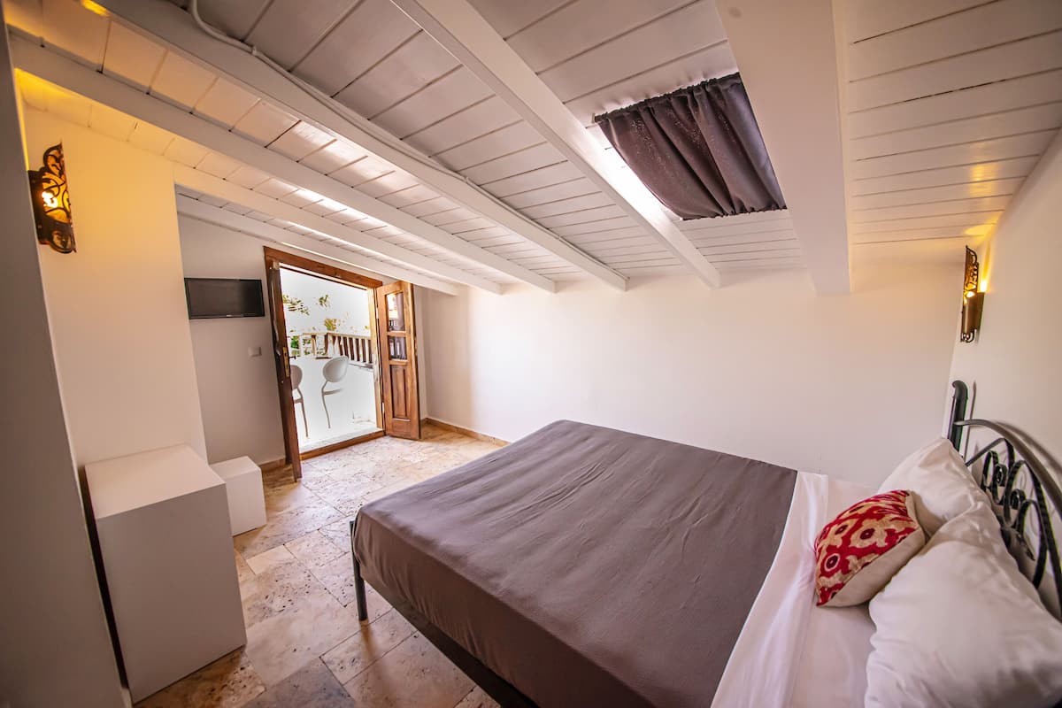 A bedroom with a bed, a small bedside table, a wall-mounted TV, and a window with curtains.