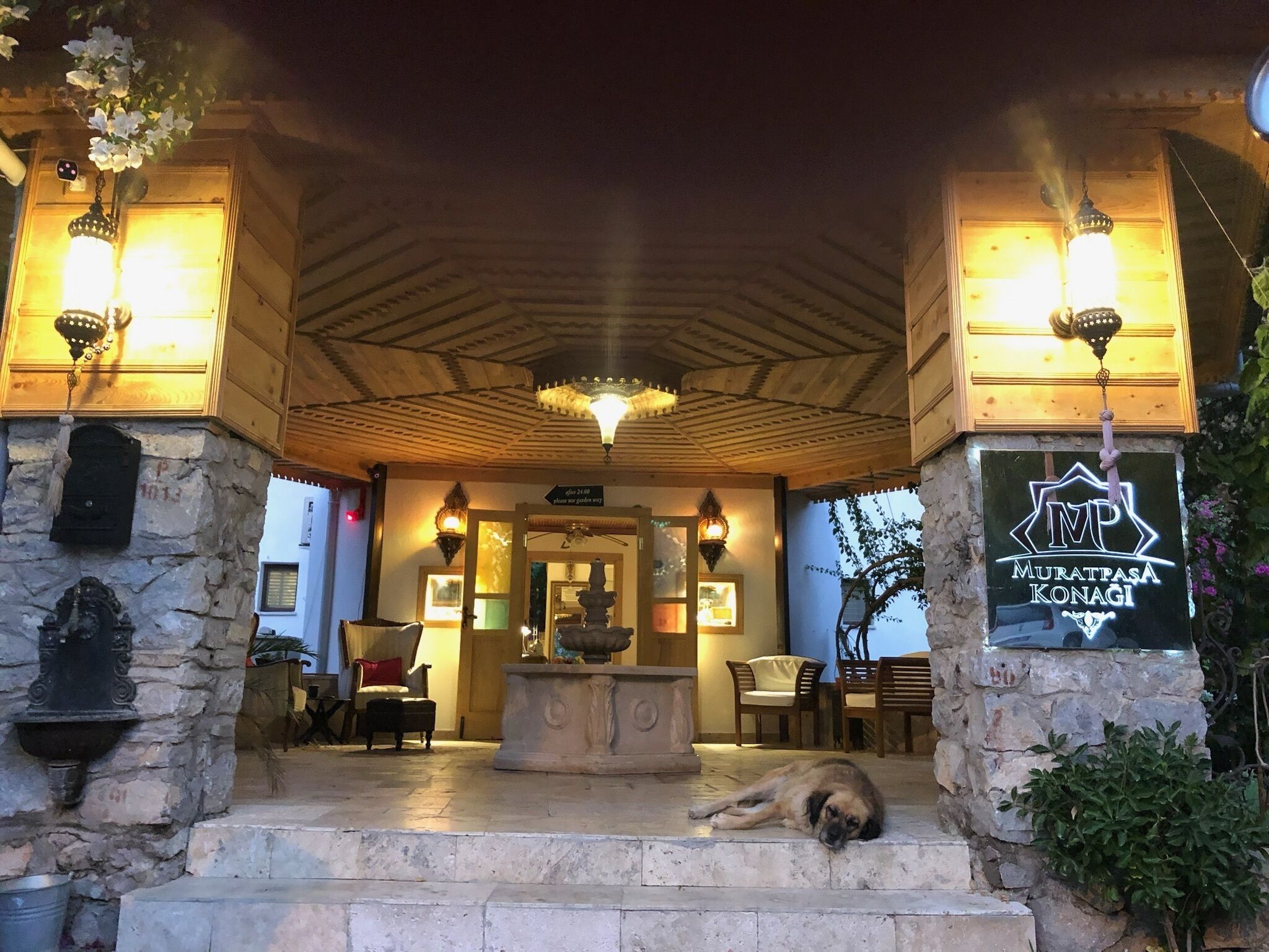 A wooden covered entrance with stone walls, a dog resting on the steps, and a sign reading "Muratpasa Konagi".