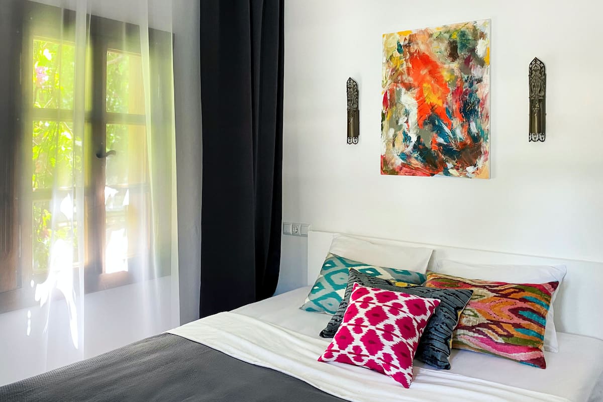 A bedroom with a bed, a window with sheer curtains, and a colorful abstract painting on the wall.