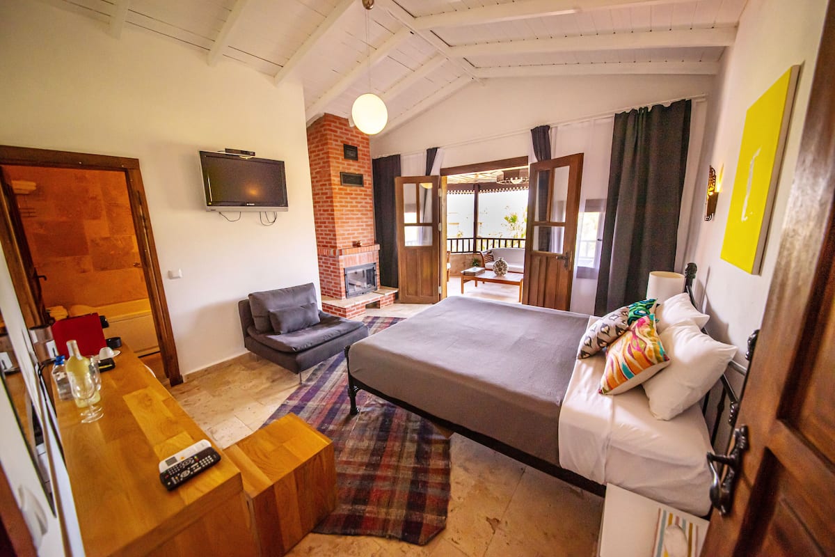 A bedroom with a bed, a TV, a fireplace, a balcony, and a wooden desk with a remote control.