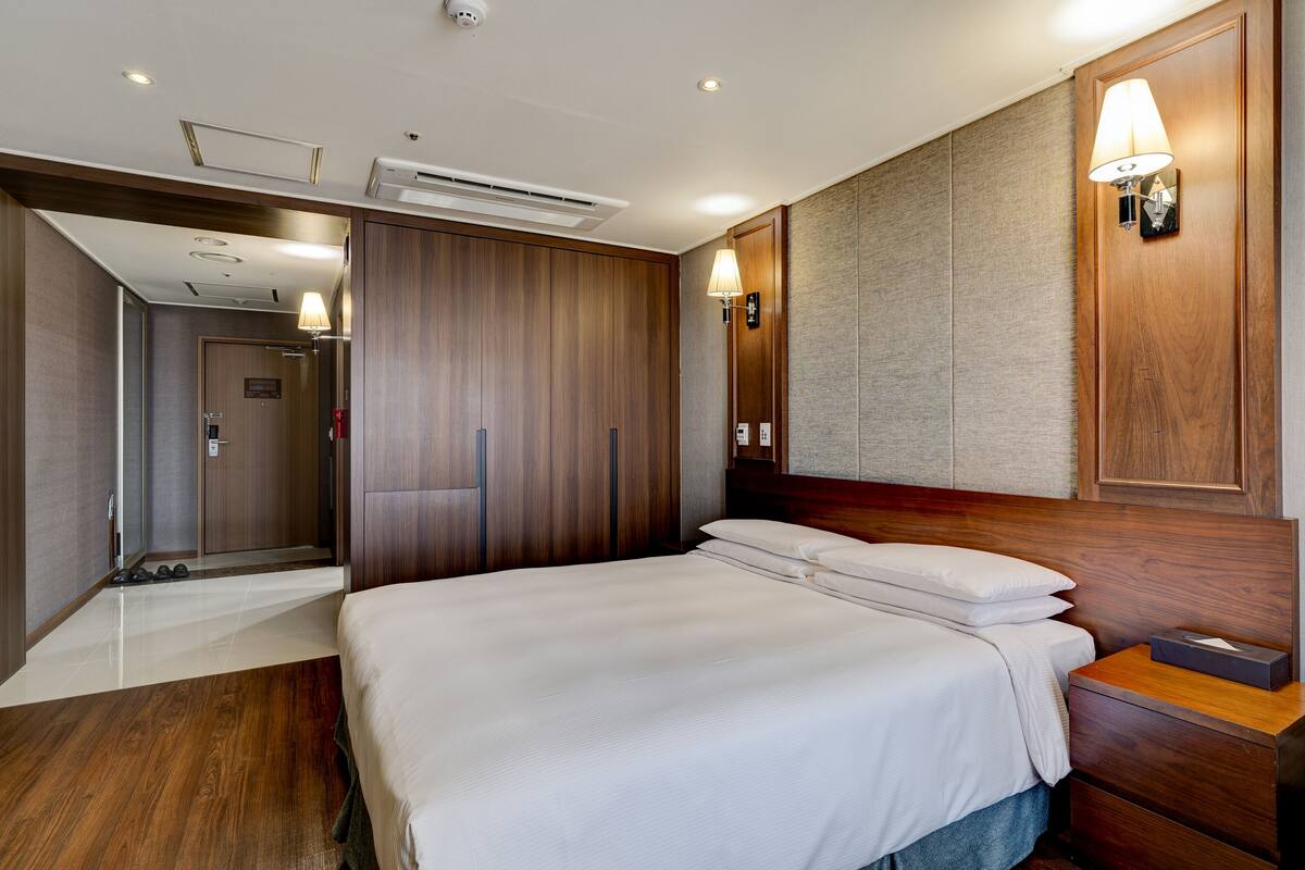 Una habitación de hotel con una cama grande, cabecera de madera, una mesita de noche, una lámpara de pared y un equipo de aire acondicionado.