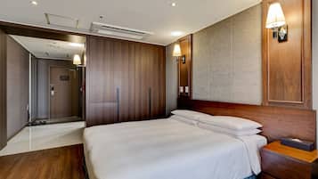 A hotel room with a large bed, wooden headboard, a nightstand, a wall-mounted lamp, and an air conditioning unit.