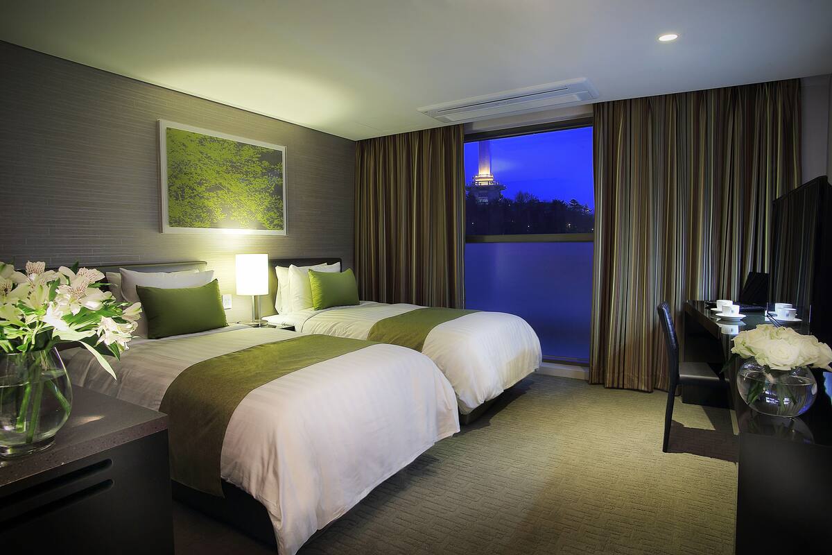 A hotel room with two beds, a desk, a chair, a TV, and a view of a cityscape at dusk.