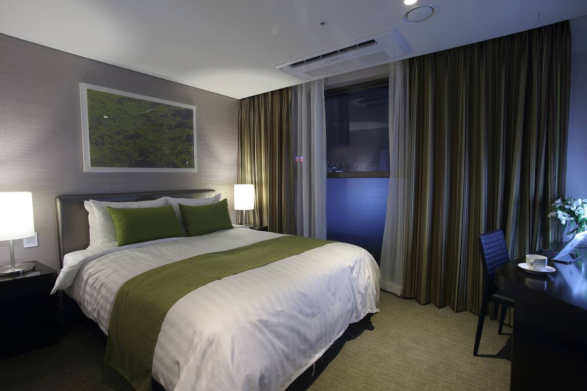 A hotel room with a large bed, a desk with a laptop, and a view of the city at night.