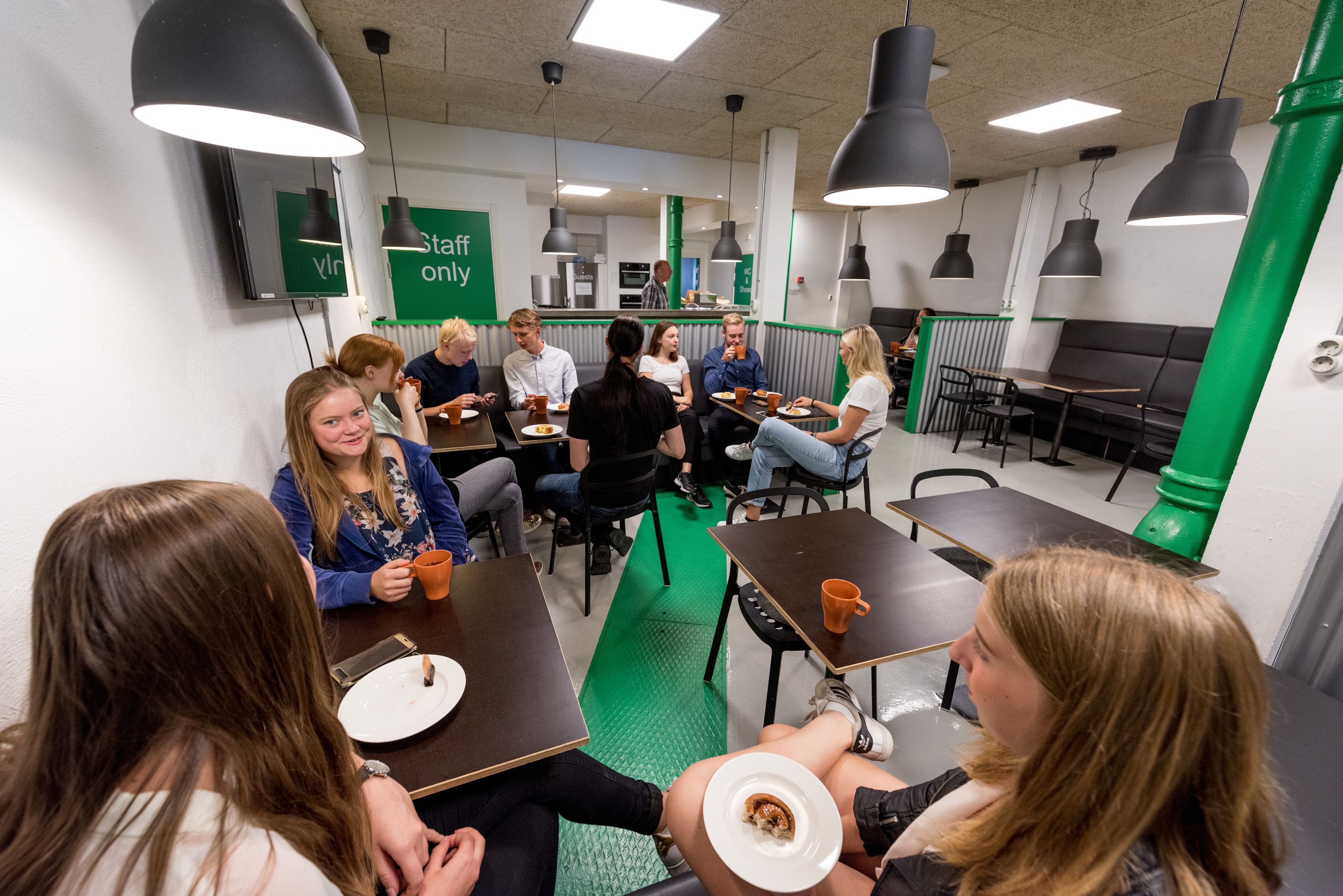 Un grupo de personas sentadas en mesas en un comedor con piso verde e iluminación colgante negra.