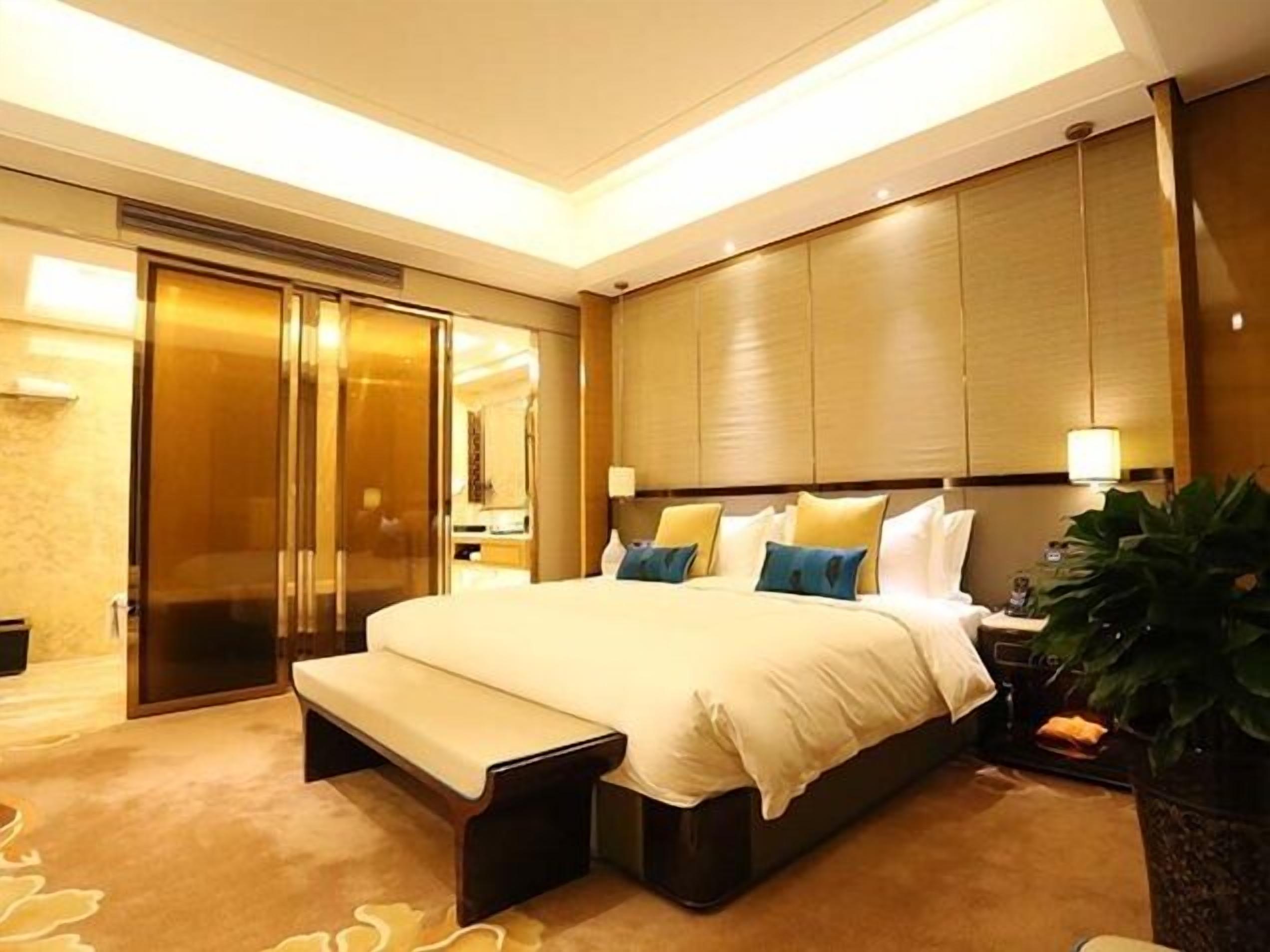 A hotel room with a large bed, a bench, a wardrobe, and a potted plant.