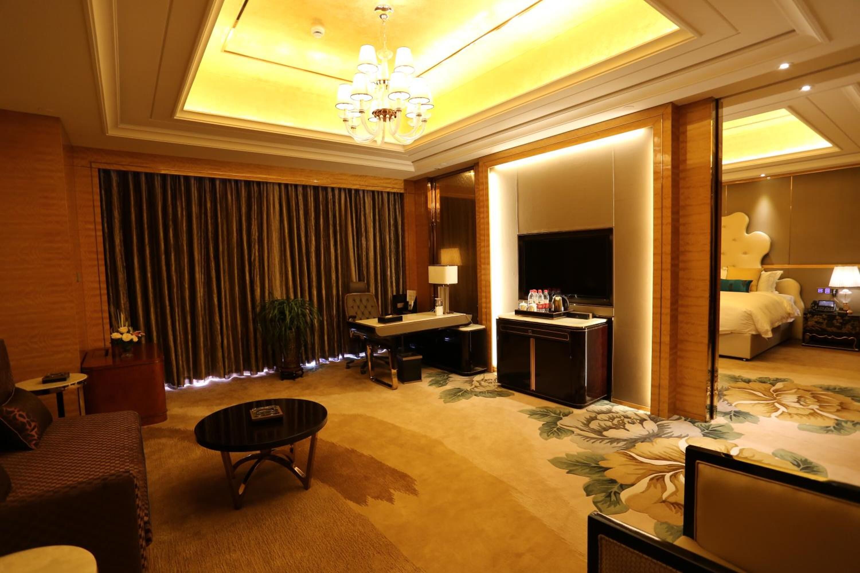 Habitación de hotel con una cama grande, televisión, escritorio con lámpara, mesa de centro y un amplio ventanal con cortinas.