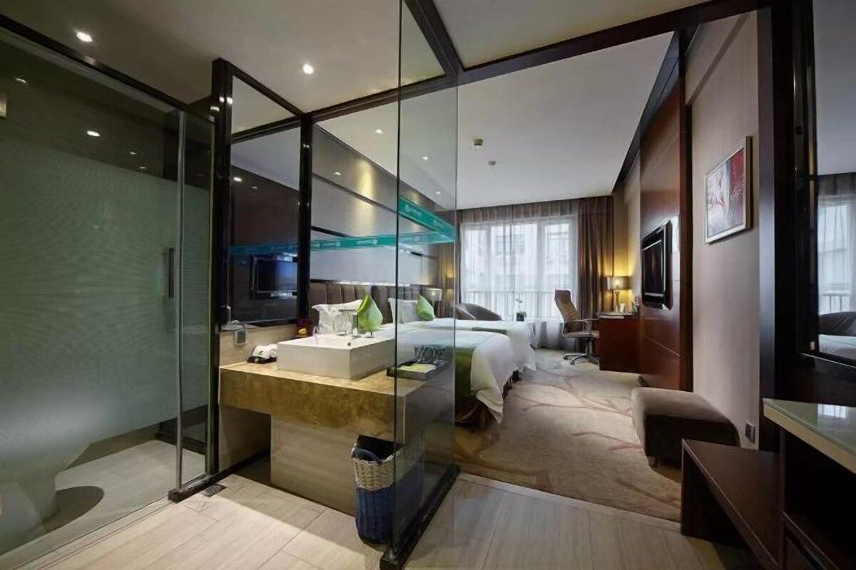 A modern hotel room with a glass partition, a bed, a desk with a computer, and a television.
