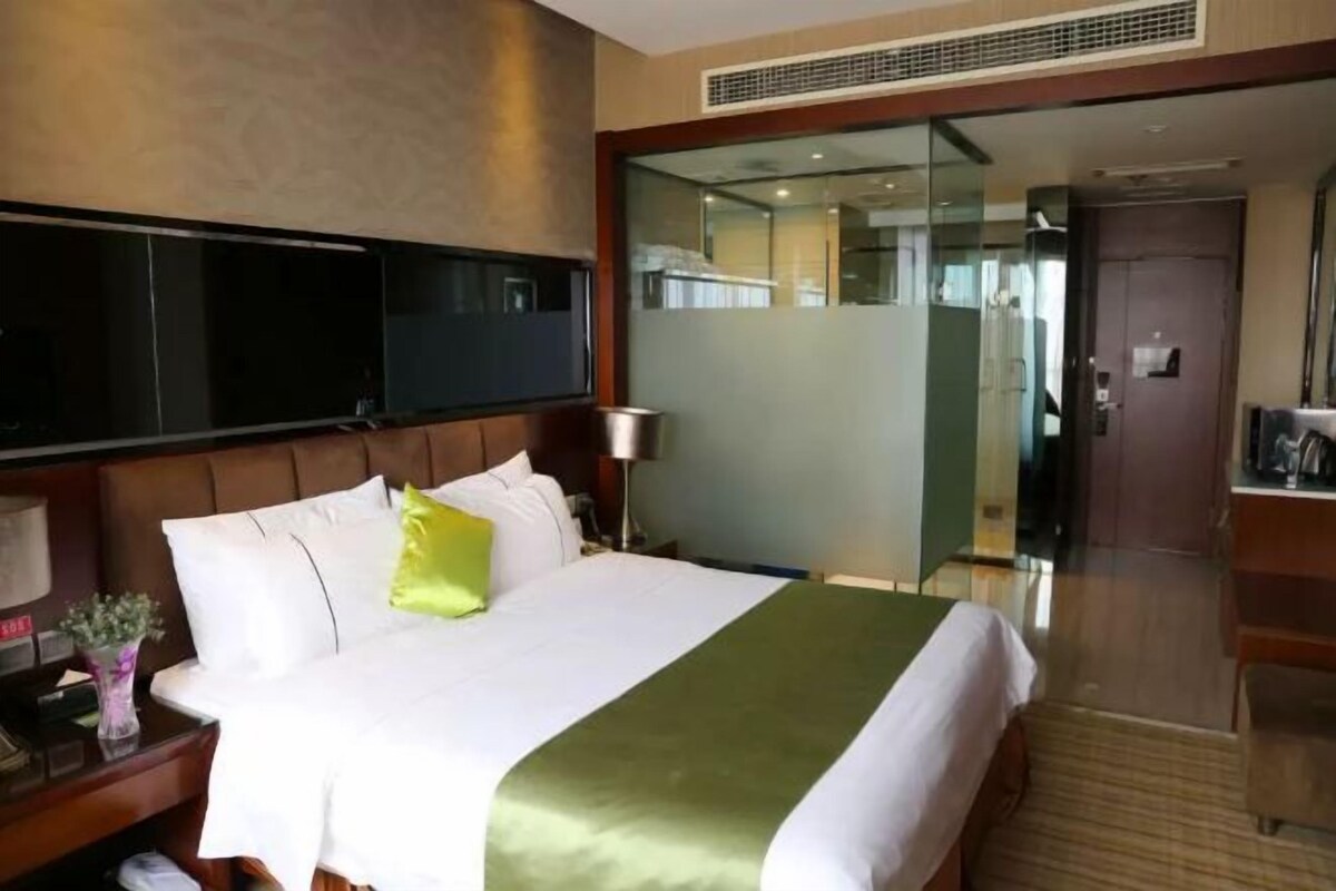 A hotel room with a large bed, a flat-screen TV, a bedside table with a lamp, and a glass door leading to a bathroom.