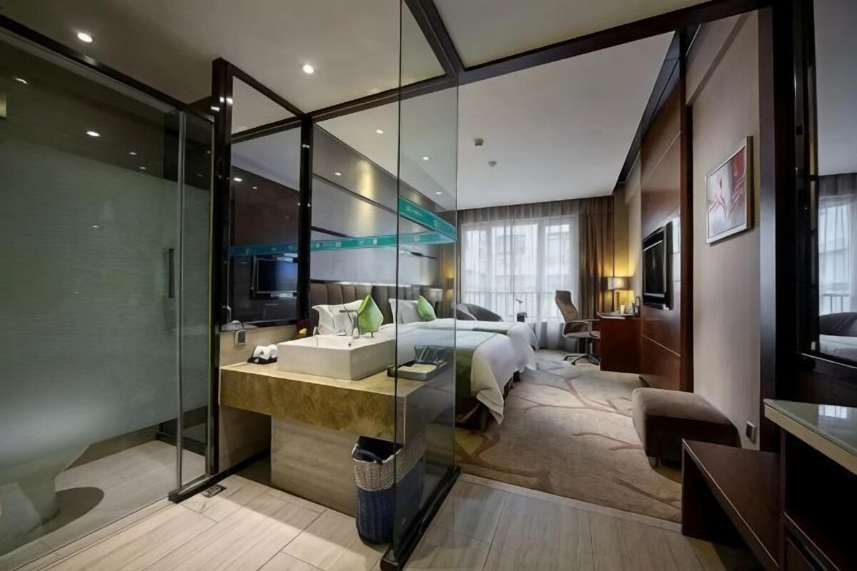 A modern hotel room with a glass partition, a bed, a desk with a computer, and a television.