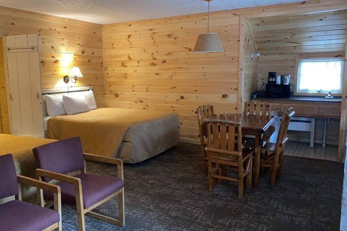A cozy cabin-style room with a bed, two chairs, a dining table, and a kitchenette.