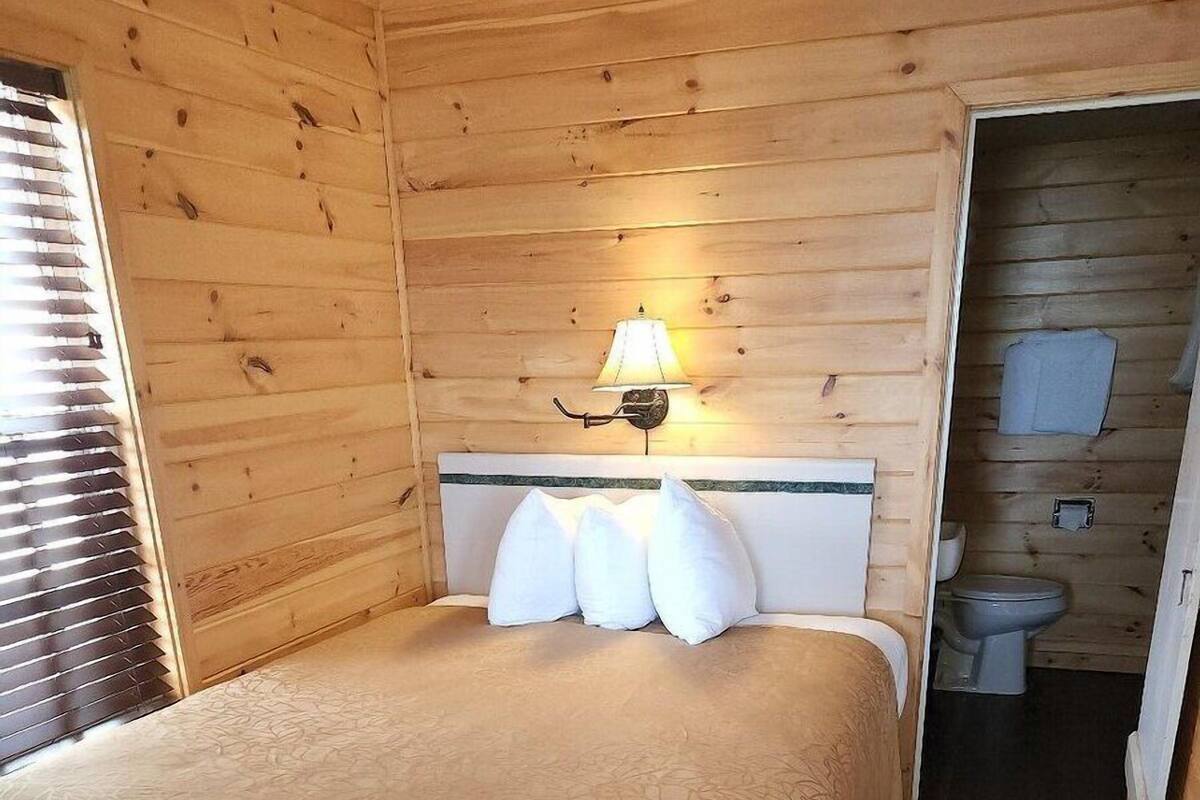 A wooden-paneled room with a bed, a nightstand, a lamp, and a bathroom visible through an open door.